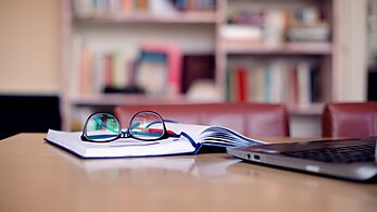Reading glasses on top of an open book with a laptop on top of a desk
