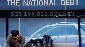 Homeless man sits at a bus stop below a sign displaying the national debt