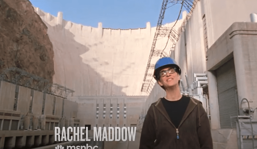 Rachel Maddow stands in front of the Hoover Dam wearing a blue helmet
