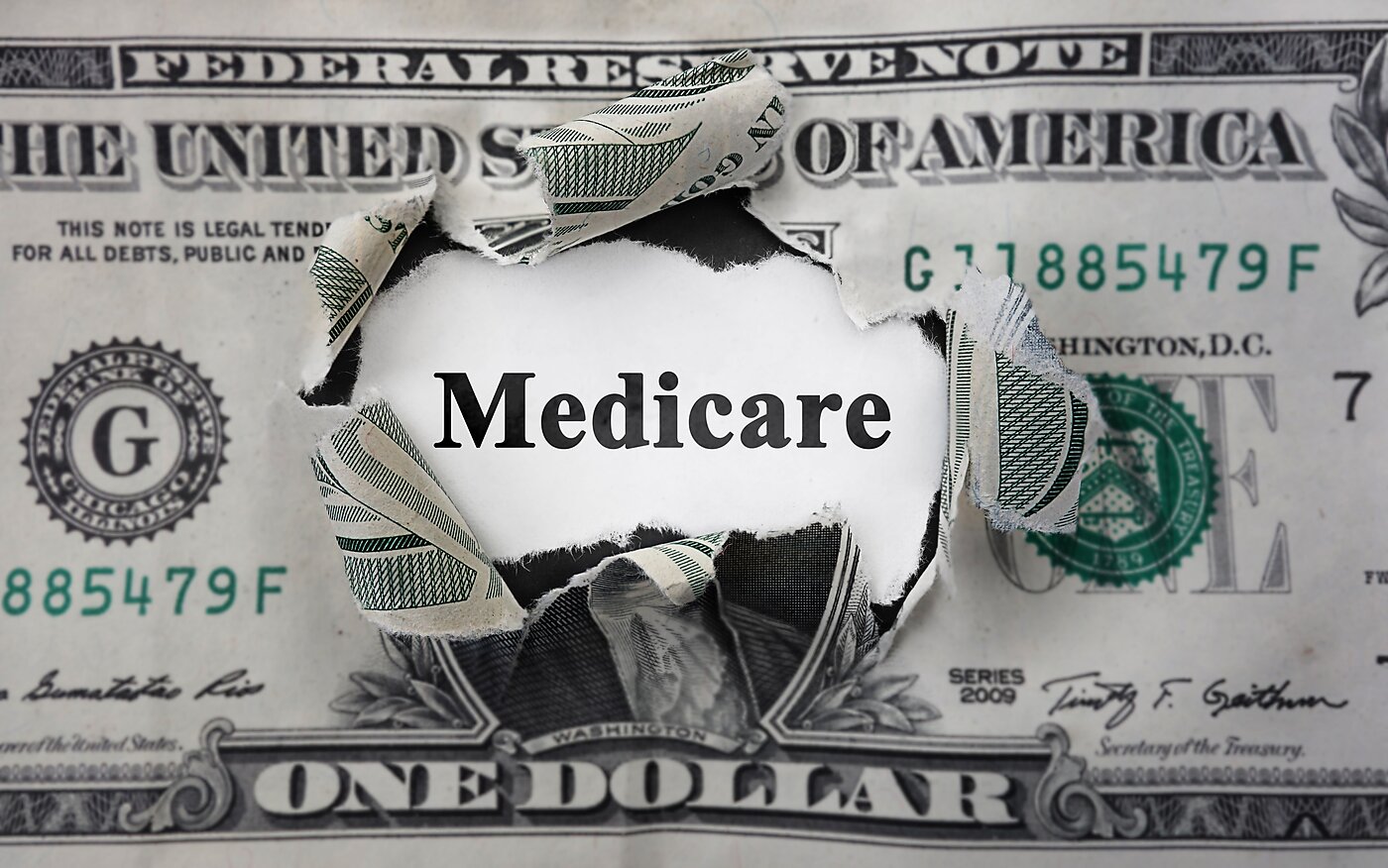 “Torn U.S. dollar bill with the word ‘Medicare’ revealed through the rip.”